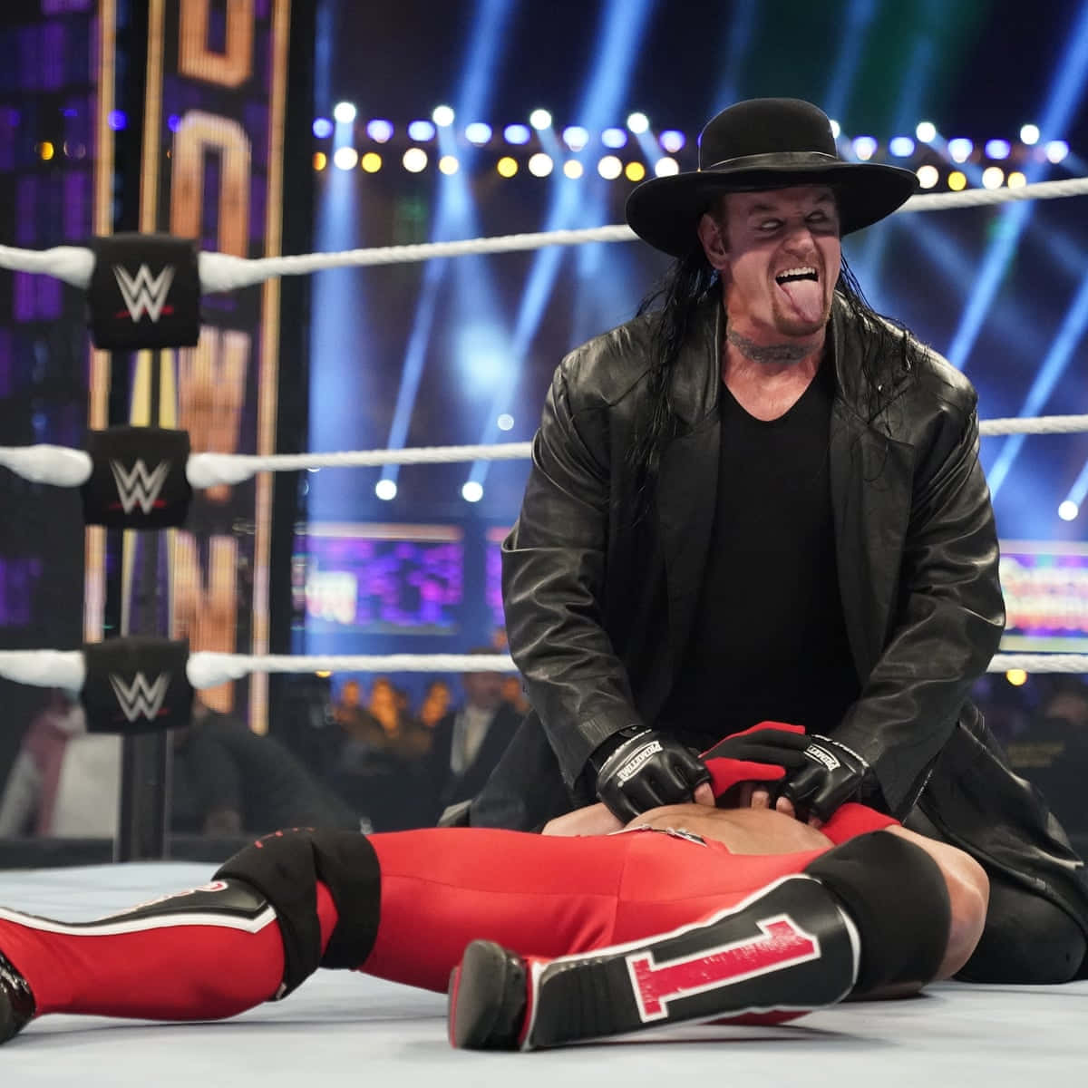  壁紙 — The Undertaker 身穿黑色皮革和帽子，在燈光通明的摔角場上對著一名倒下的紅色摔角手瘋狂大笑。