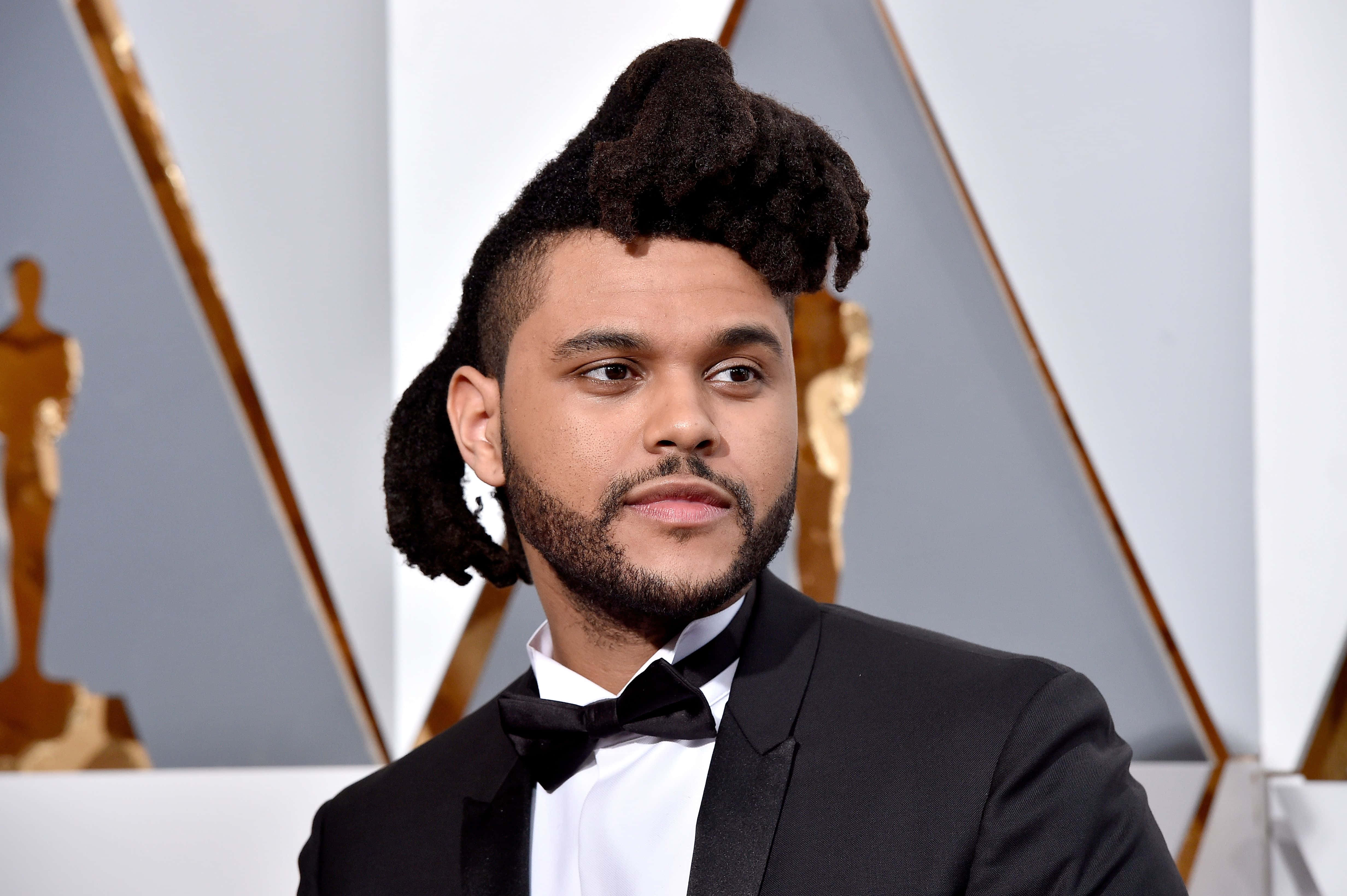 The Weeknd After Hours 桌布 壁紙 — The Weeknd 身穿黑色燕尾服配蝴蝶結，以金色奧斯卡獎座和幾何銀色面板為背景擺姿勢。