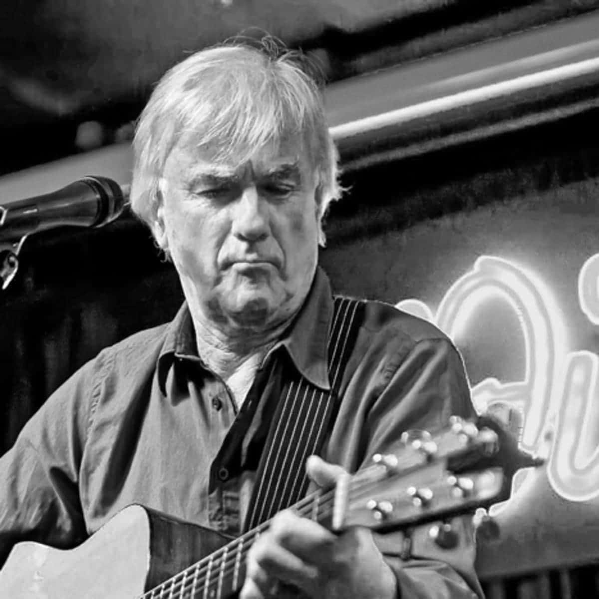 The Yardbirds 鼓手 Jim McCarty 老年時演奏 壁紙 — 一個黑白肖像，一個男人在發光的霓虹燈牌下激烈地彈奏木結他。
