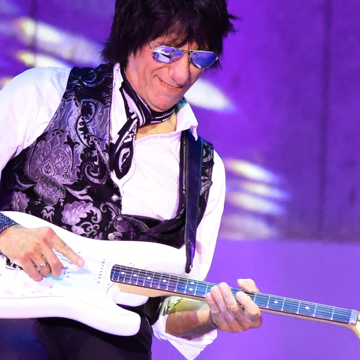The Yardbirds 前成員 Jeff Beck 吉他即興演出 壁紙 — 一位搖滾結他手戴着紫色太陽眼鏡和圖案背心，在充滿活力的紫色舞台燈光下熱情地彈奏白色電結他。
