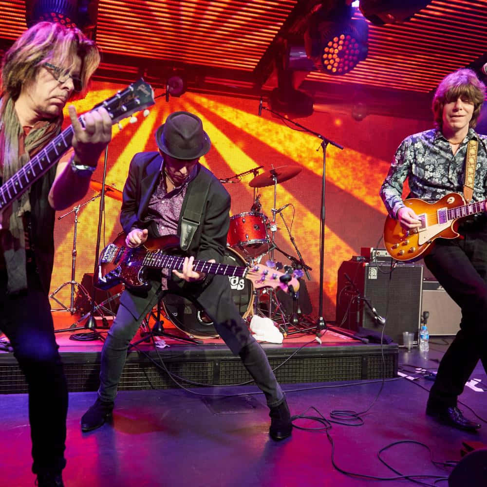 The Yardbirds 現役成員 Johnny A、Kenny Aaronson 與 John Idan 演出照 壁紙 — 三位音樂家在充滿活力的舞台上，在紅色和橙色聚光燈下激烈地演奏電結他。