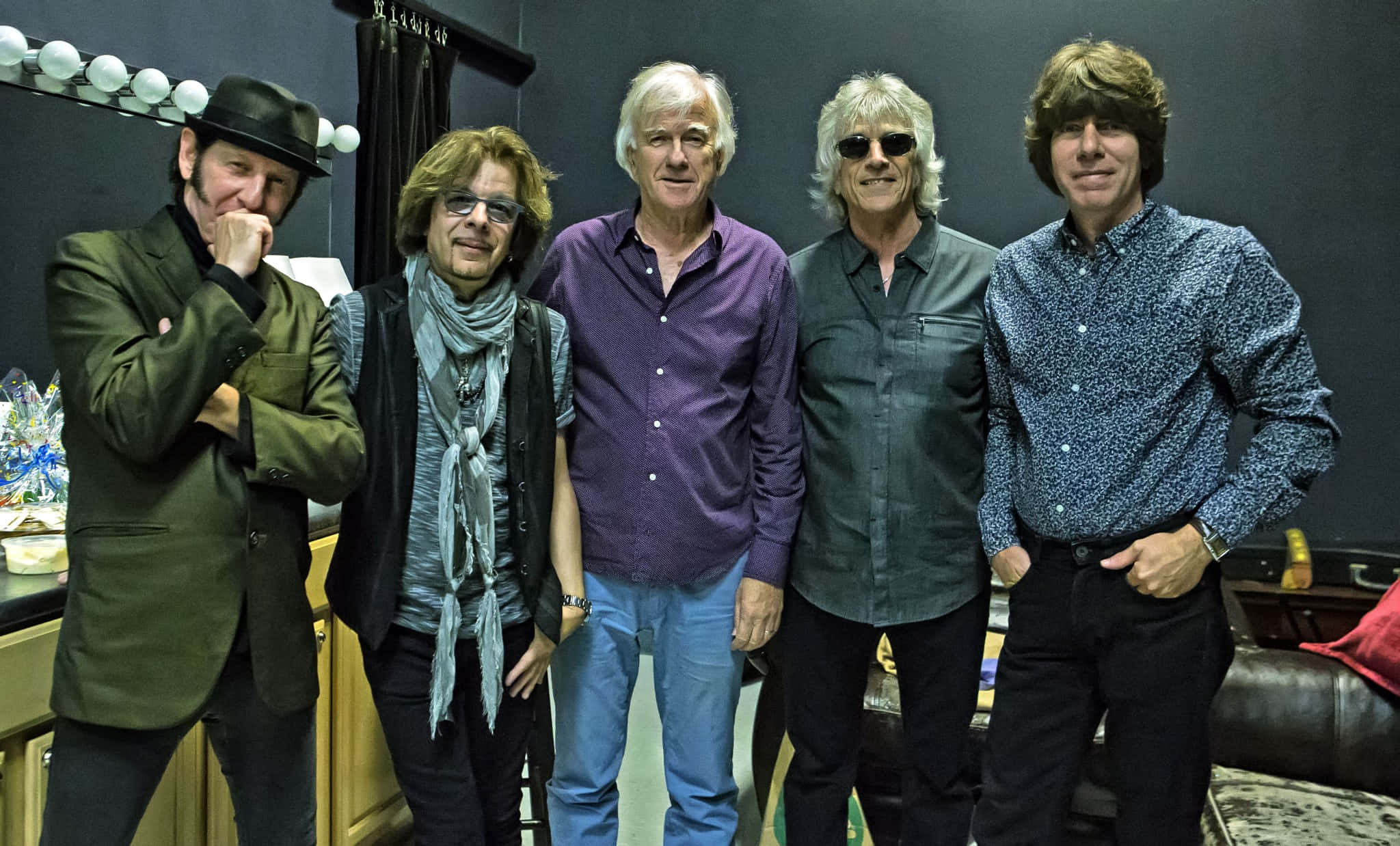 The Yardbirds 現役成員與創始鼓手吉姆·麥卡蒂 壁紙 — 五位身穿時尚休閒服的男士在後台化妝間自信地擺姿勢，房間內有化妝鏡和皮革沙發。