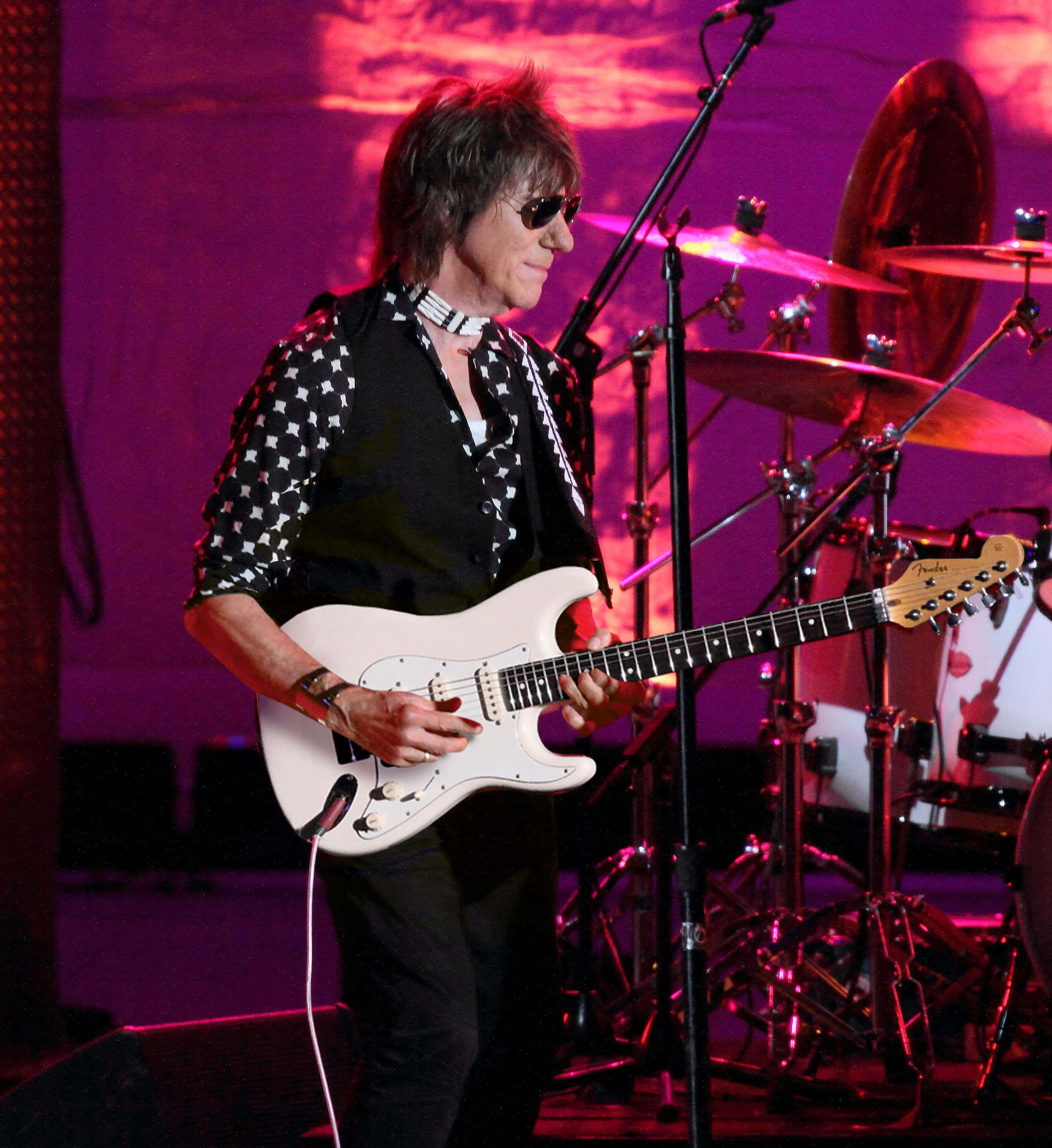 The Yardbirds 艾夫·貝克 2022 英國巡演 壁紙 — 一位搖滾音樂家在舞台上彈奏白色電結他，背後有鼓組，舞台燈光呈現鮮豔的粉紅色和紫色。