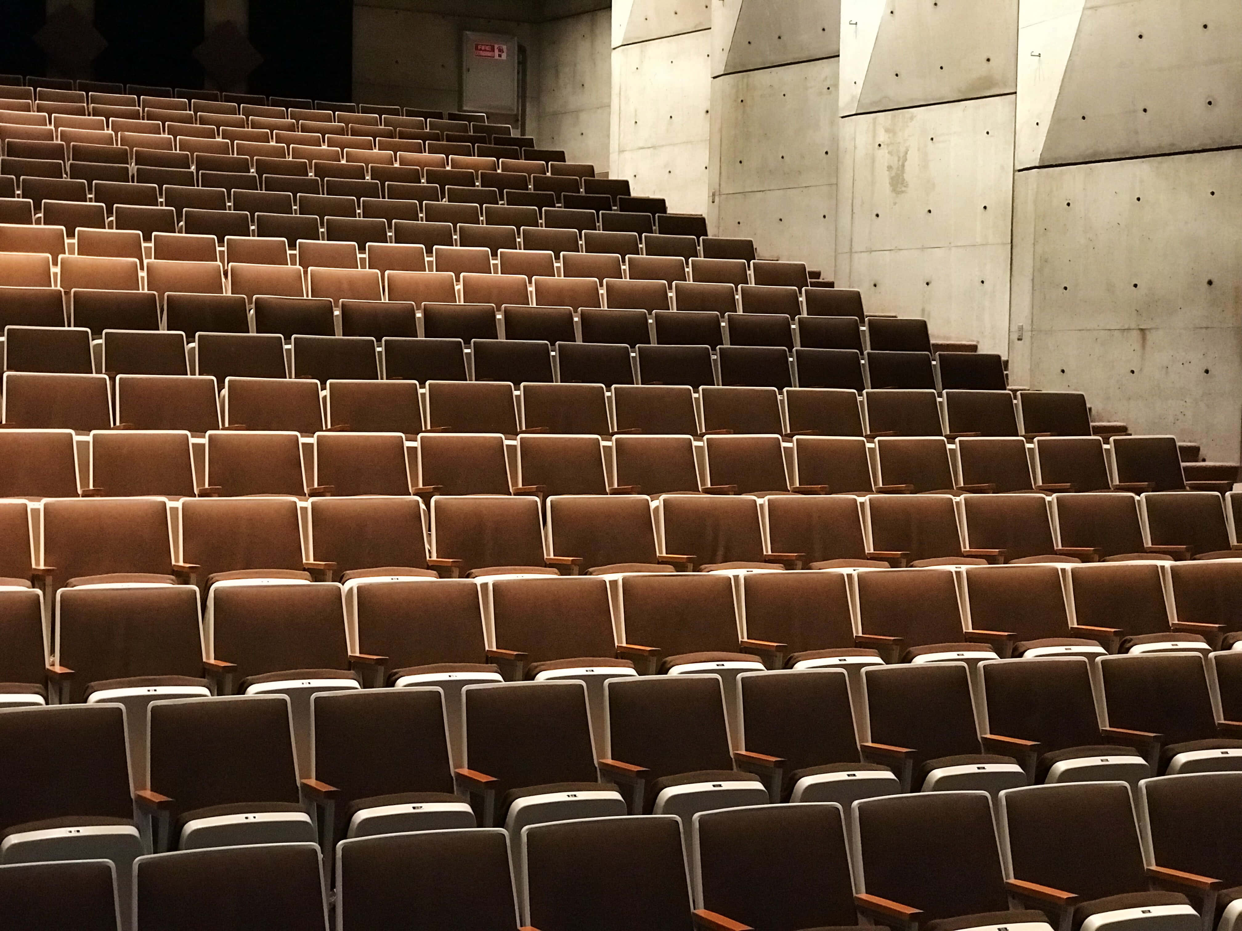 法國 Theatre Melchior 電影院背景 壁紙 — 一排排空的棕色劇院座位在溫暖的環境光線下向混凝土牆上升。