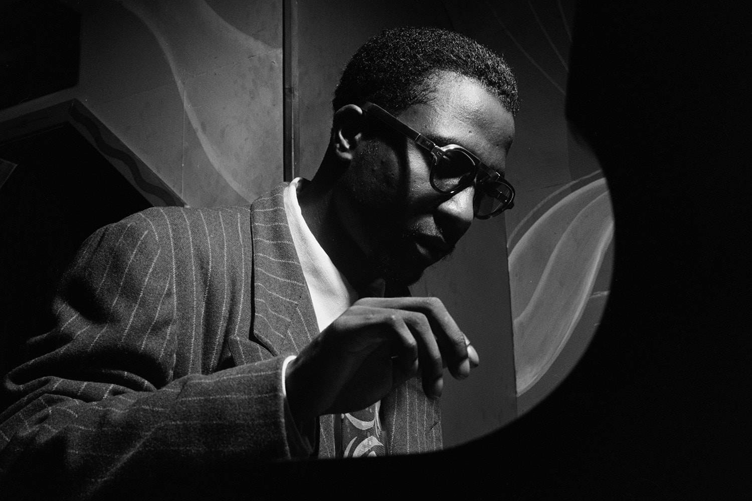 Thelonious Monk 單色鋼琴演奏 壁紙 — 一名穿著細條紋西裝和眼鏡的男子，在戲劇性的黑白燈光下透過圓形窗戶凝視。
