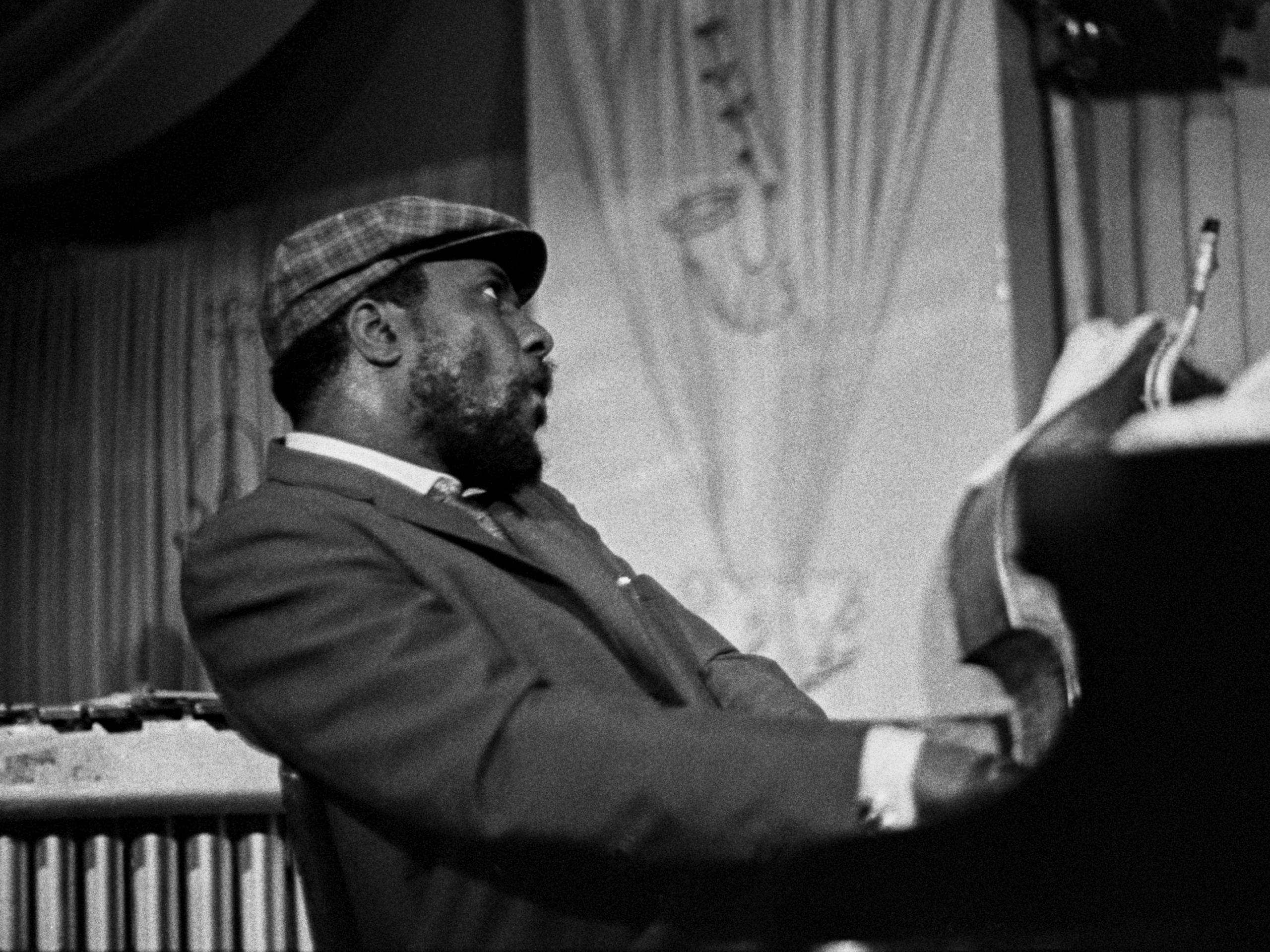 Thelonious Monk戴帽彈琴 壁紙 — 一位戴格仔帽、身穿西裝的爵士鋼琴家在戲劇性燈光下於舞台上熱情演奏。