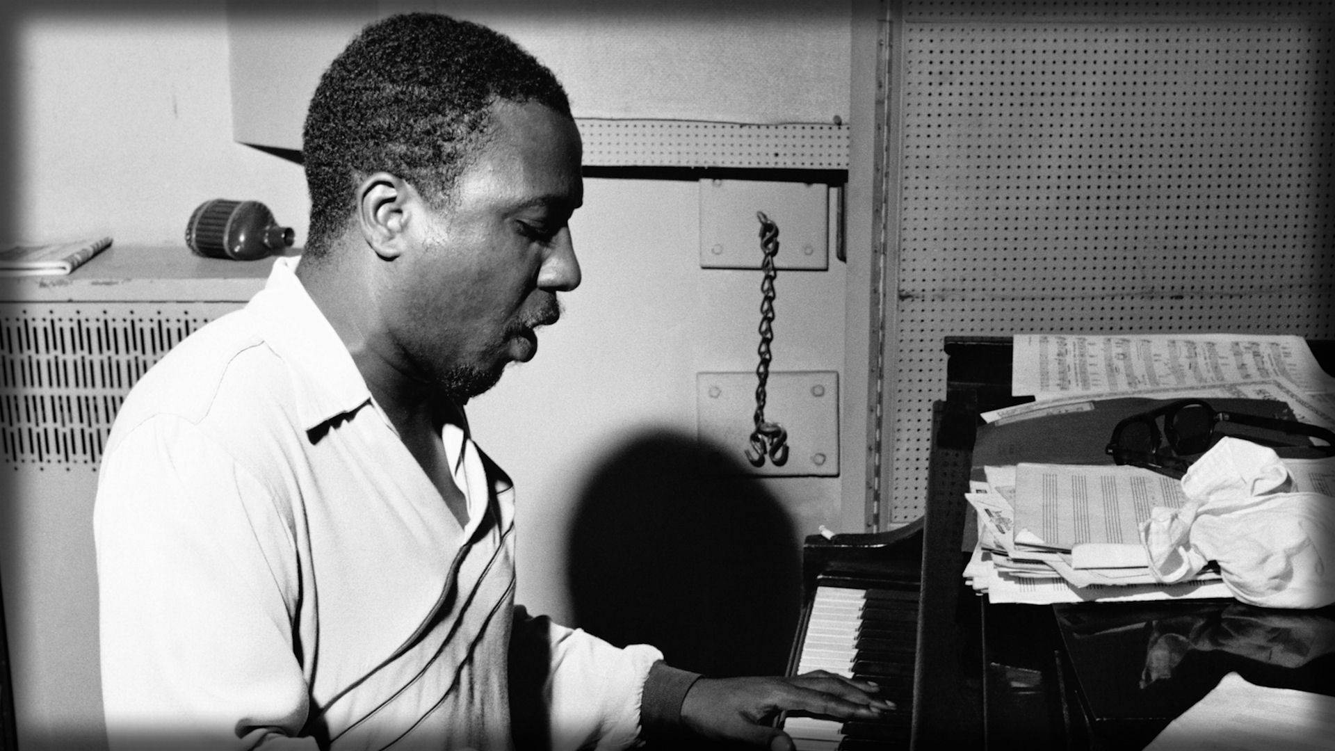 Thelonious Monk 聚焦創作音樂 壁紙 — 一張黑白照片，一名男子在雜亂的錄音室裡專注地彈鋼琴，周圍有樂譜和牆上掛著的鏈條。