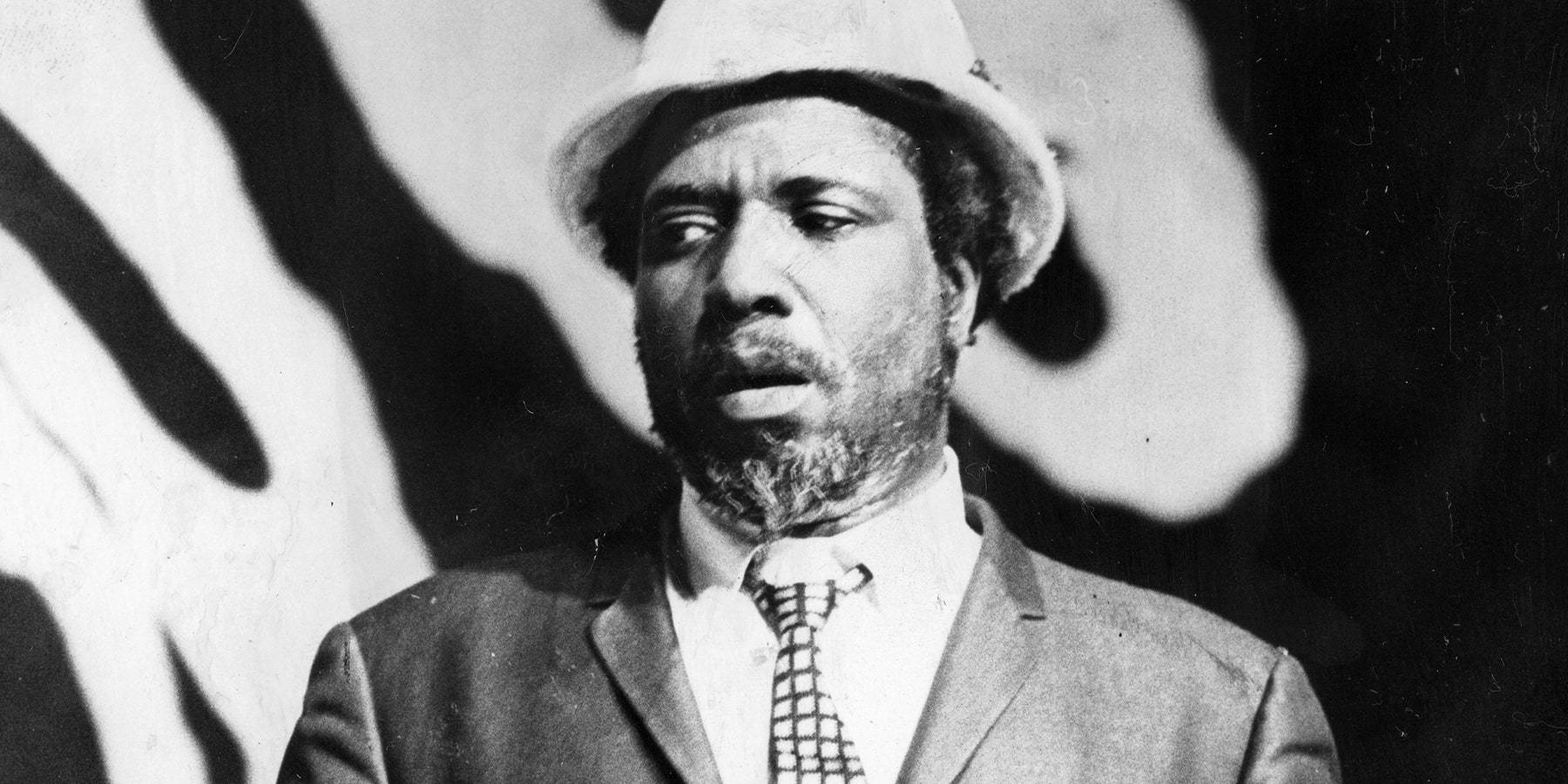 Thelonious Monk 沉思時刻 壁紙 — 一個穿著西裝和軟呢帽的男人，在圖案背景前向上看，表情嚴肅的黑白肖像。