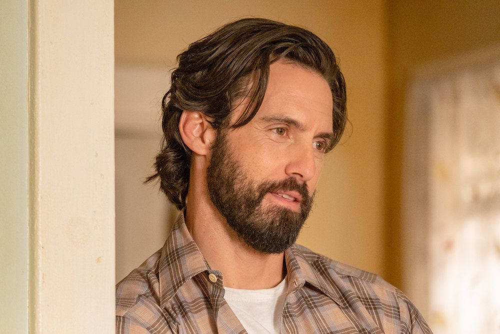 「傑克·皮爾森——《This Is US》的靈魂人物」 壁紙 — 一位留著深色波浪髮和鬍鬚的男子，在室內溫暖燈光下穿著格仔恤衫。