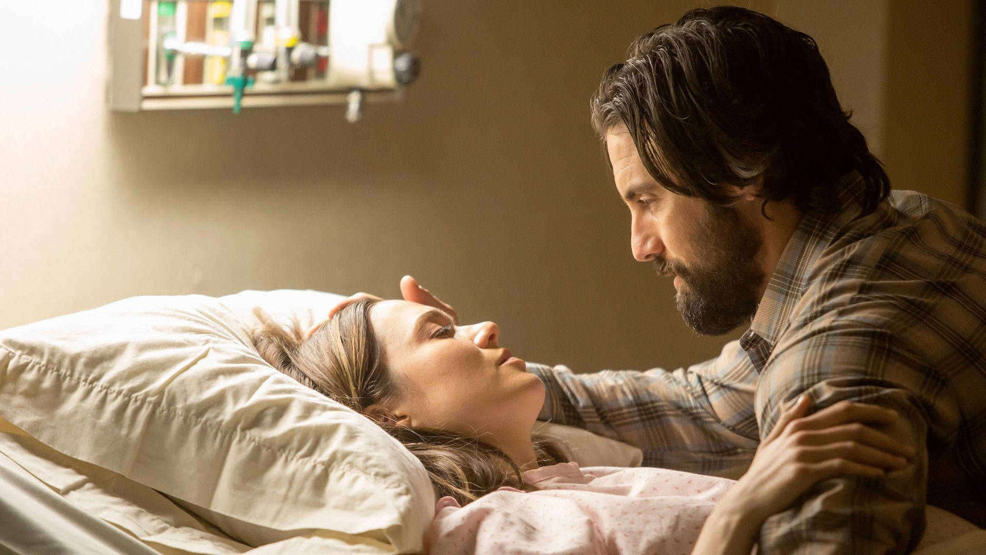《This Is US》傑克與麗貝卡 壁紙 — 一名男子輕柔地觸摸躺在醫院病床上的女子的額頭，柔和的自然光從窗戶灑入。