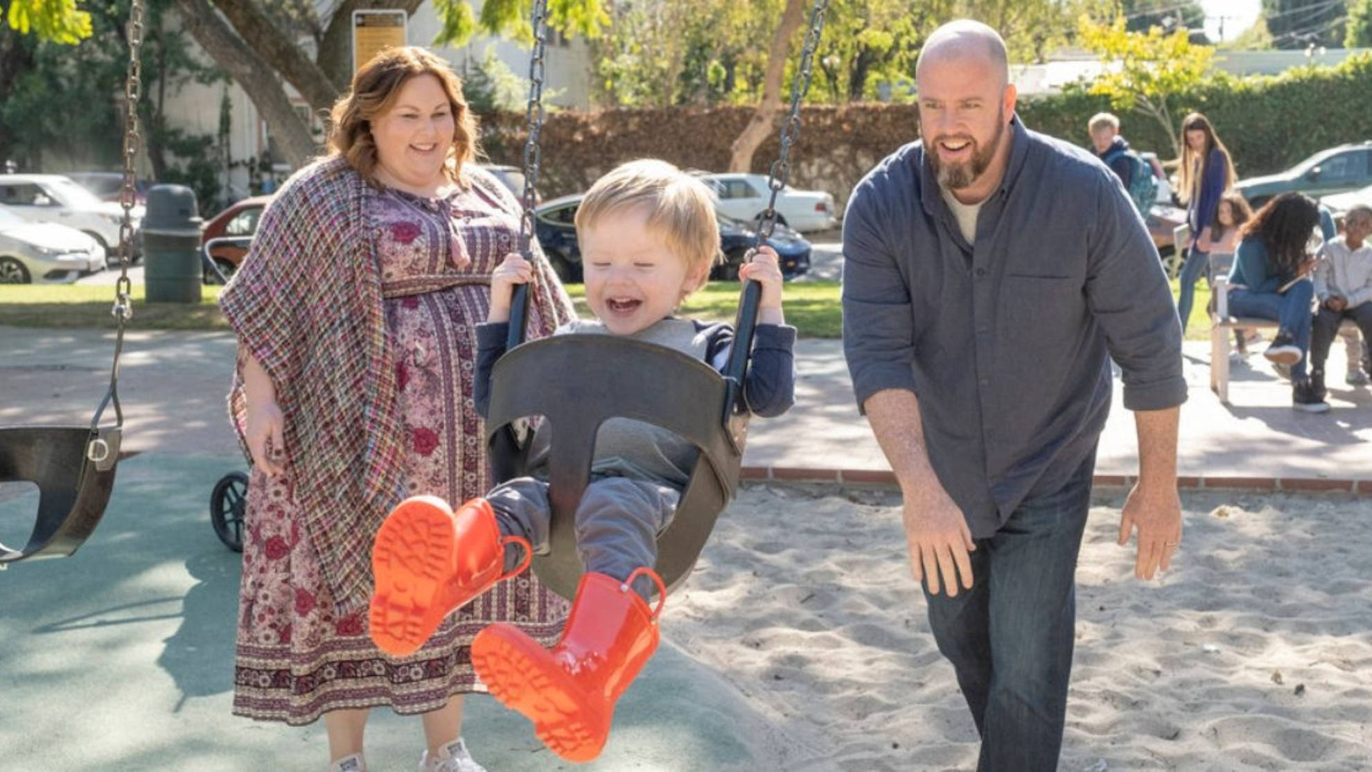 《This Is US》凱特與托比 壁紙 — 在陽光明媚的遊樂場上，一個穿著鮮紅色靴子、大笑的孩子在兩個成人之間盪鞦韆的快樂家庭時刻。