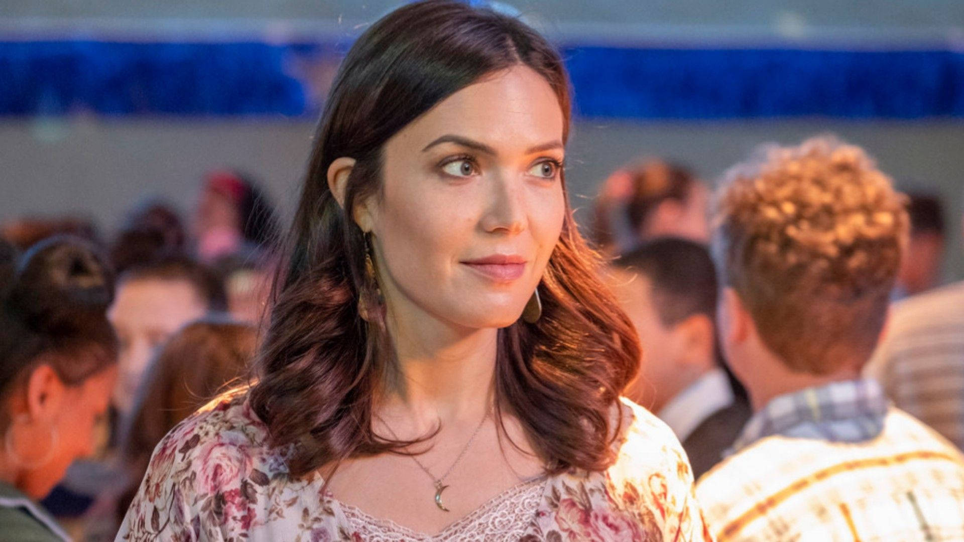 《This Is US》曼迪·摩爾 壁紙 — 一位擁有波浪棕色頭髮和花卉襯衫的女性，在擁擠的社交場合中若有所思地看著鏡頭外的某人。