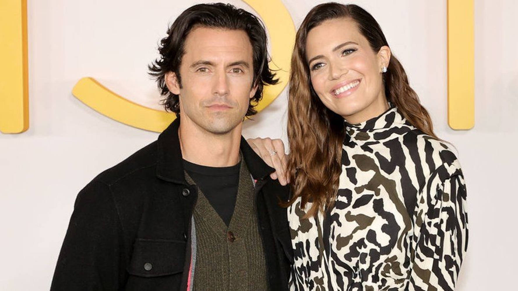 《This Is US》虛構情侶角色 壁紙 — 一男一女在一個有亮黃色標誌背景的活動中一起擺姿勢。