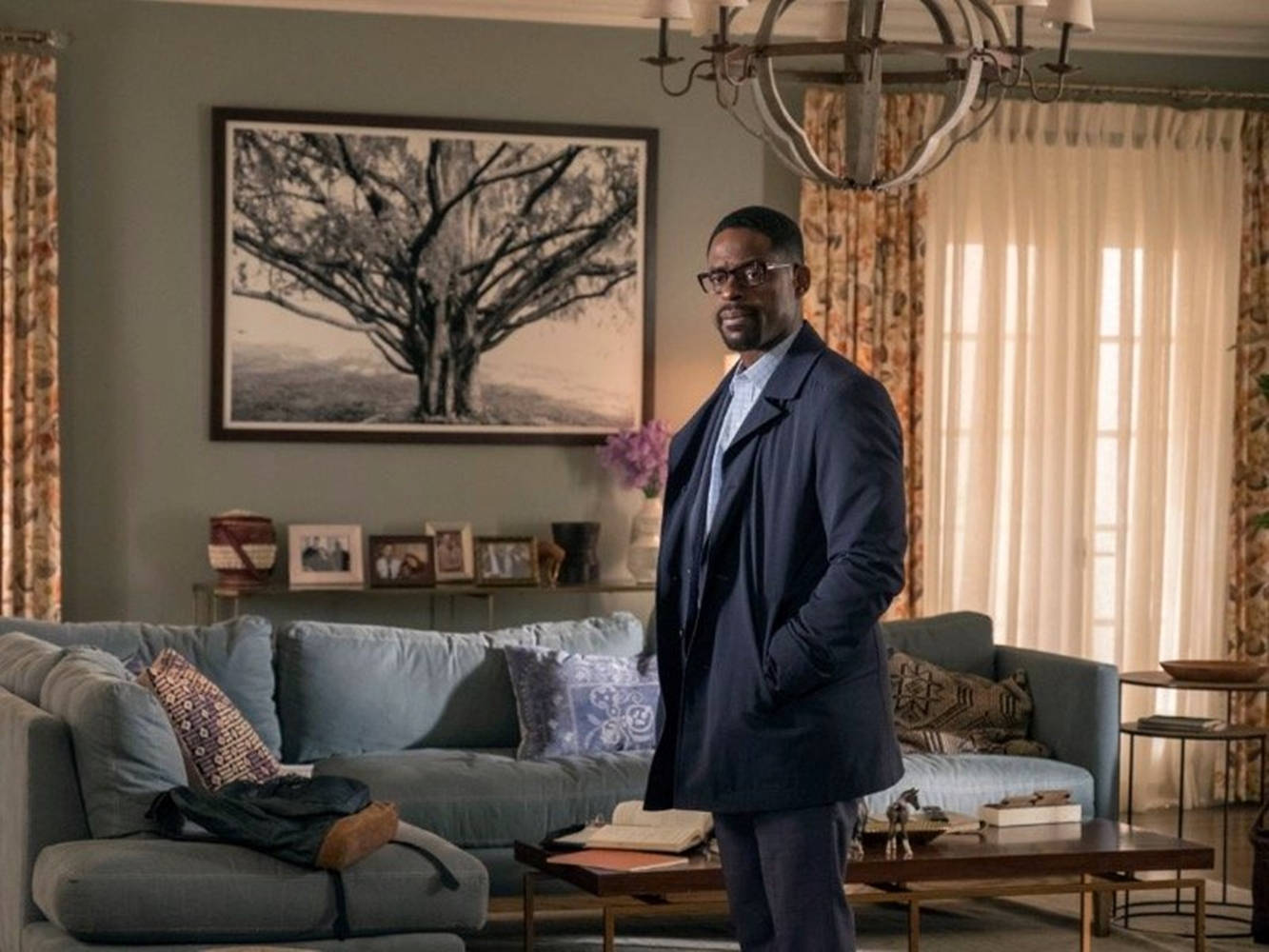 《This Is US》角色蘭道爾·裴森 壁紙 — 一名身穿深色西裝的男子自信地站在陽光充足的客廳中，牆上掛著一幅大型樹木藝術品。