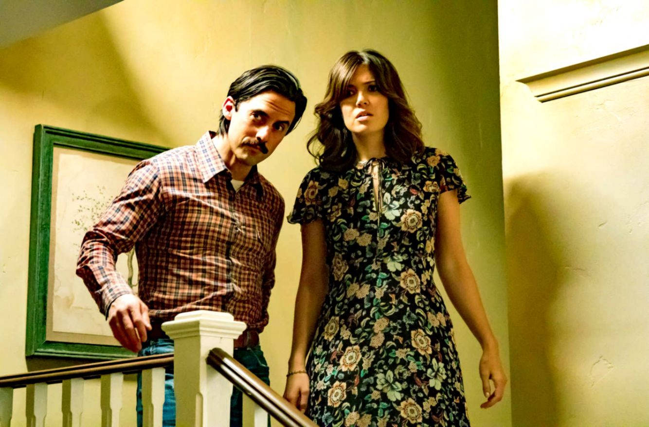 《This Is US》NBC原創劇集 壁紙 — 一位留鬍子的男人和一位穿著花卉裙的女人站在樓梯上，樓梯位於一個燈光溫暖的走廊，牆上掛著一幅裱框的畫。