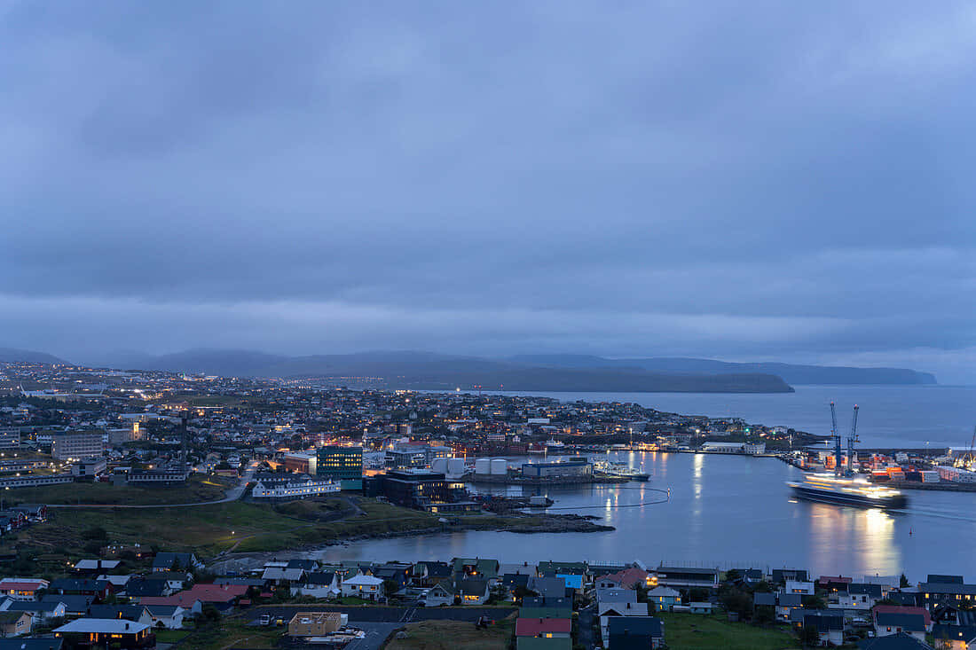 Thorshavn 在夢幻藍天之下 壁紙 — 黃昏時分的沿海小鎮，燈光閃爍，映照在平靜的水面和遠山之上，天空多雲。