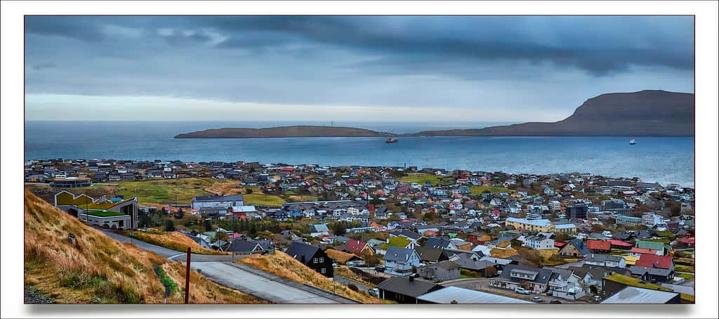 Thorshavn 城市全景與島嶼背景 壁紙 — 一個沿海小鎮在戲劇性的多雲天空下，遍佈於俯瞰平靜大海的山坡上。