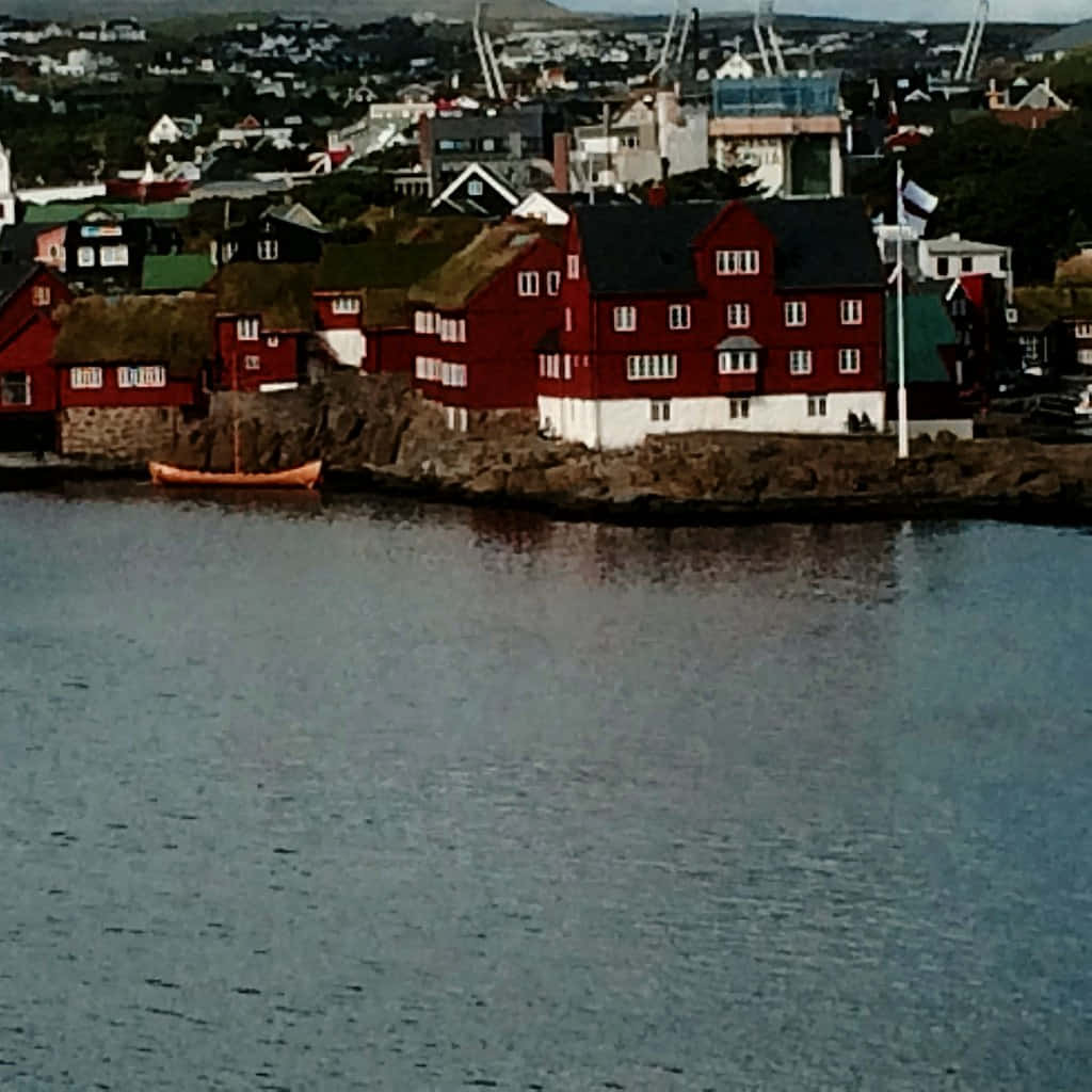 Thorshavn 海洋與 Tinganes 半島 壁紙 — 紅白相間的房屋緊貼著岩石海岸，旁邊是平靜的水面，附近漂浮著一艘橙色小船。