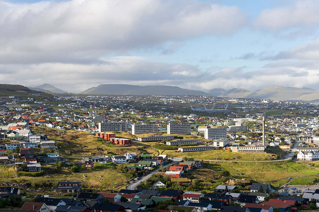 Thorshavn 的建築與房屋 壁紙 — 一個廣闊的海岸小鎮，坐落在綠色山丘中，散落著房屋和遠處的山脈，天空部分多雲。