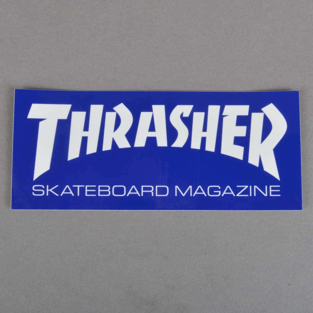 與 Thrashers 一同滑板！ 壁紙 — 一張藍色Thrasher Skateboard Magazine貼紙，帶有粗體白色文字，平放在純灰色表面上。