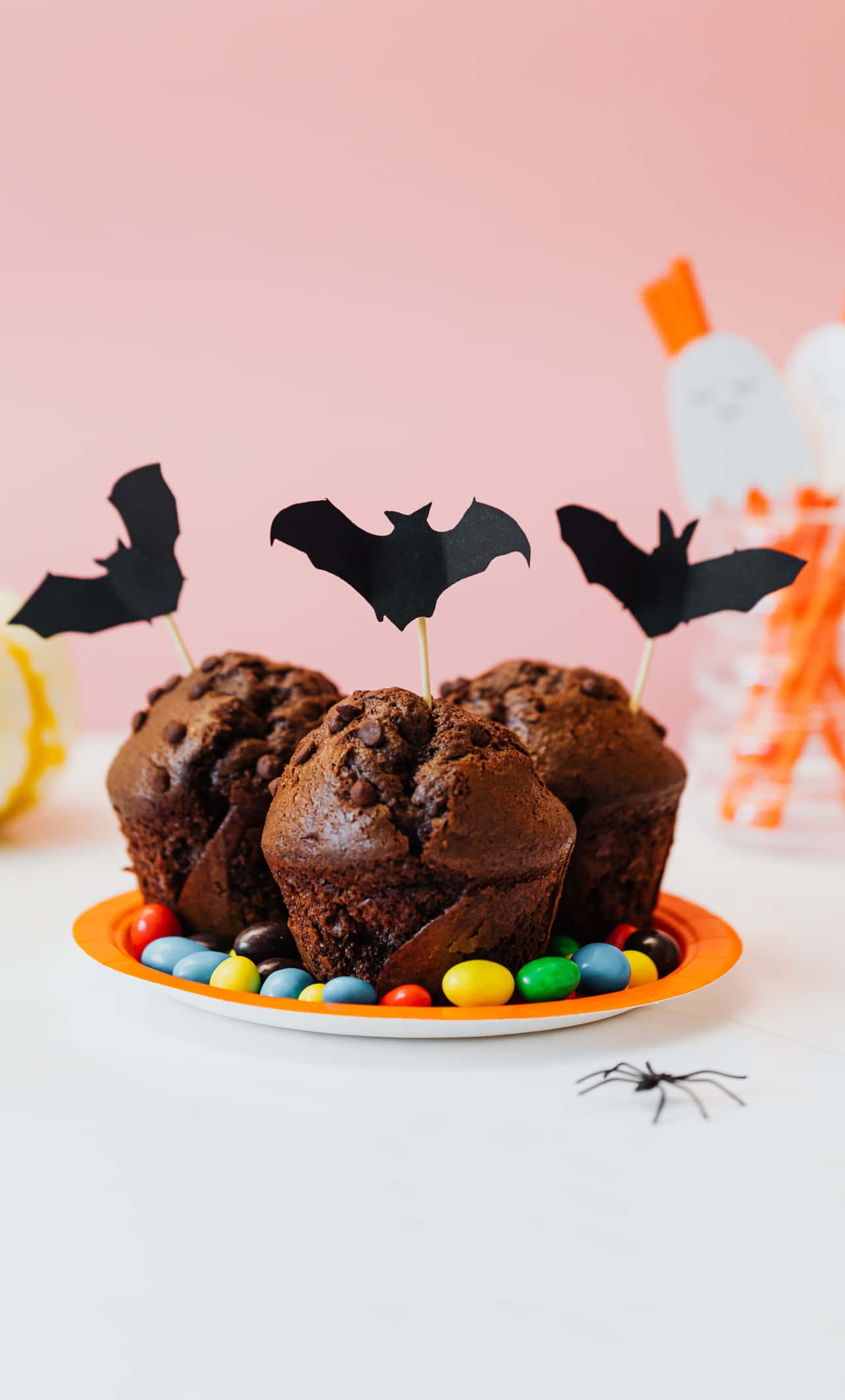 Halloween Treats 桌布 壁紙 — 三個朱古力碎鬆餅，上面插有蝙蝠裝飾棒和彩色糖果，放在粉紅色背景的碟子上。