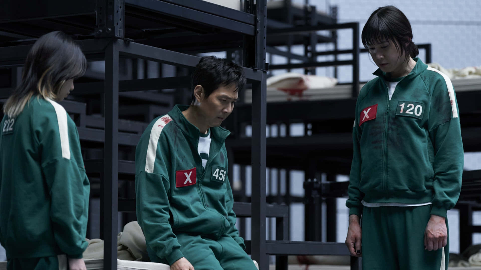  壁紙 — 三名參賽者身穿綠色號碼運動服，坐在或站在簡陋的金屬 bunk 房間，表情陰沉。