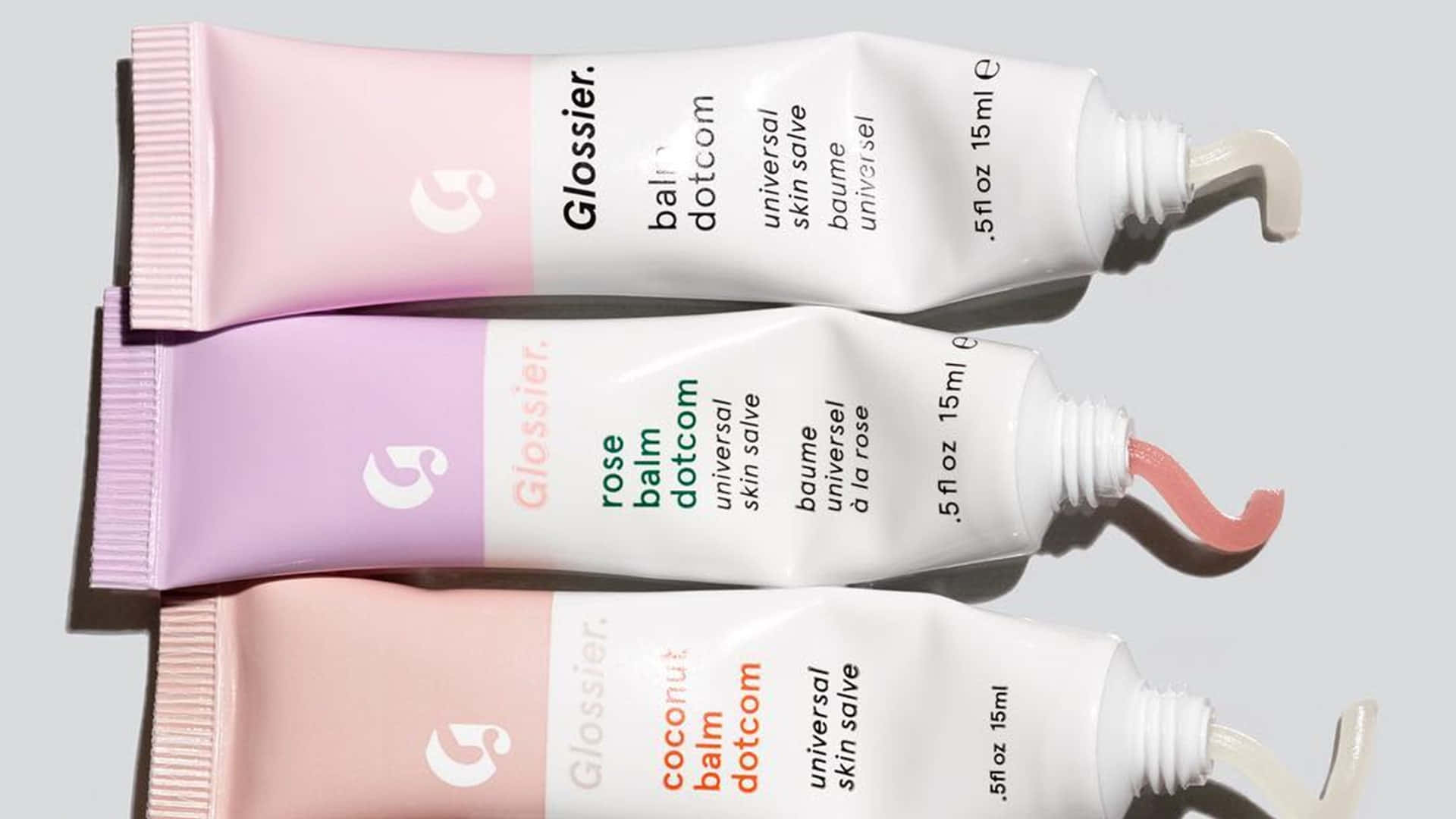  壁紙 — 三支 Glossier 潤唇膏管，分別是玫瑰、椰子及通用香味，以對角線排列，打開蓋子，放在淺灰色背景上。