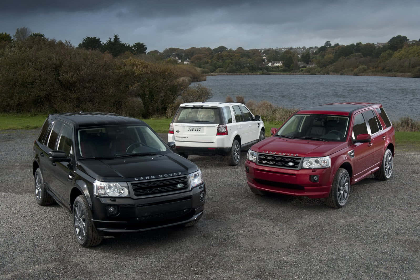 Land Rover Freelander 桌布 壁紙 — 三輛黑色、白色和紅色的Land Rover SUV停泊在湖邊的碎石地上，天空多雲。