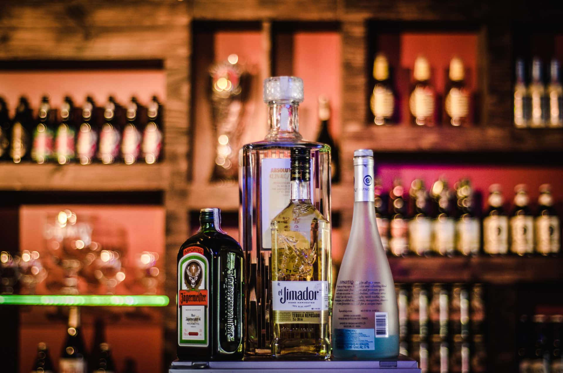  壁紙 — 三瓶酒類飲品醒目地擺放在吧枱上，背景是溫暖發光的酒瓶架。