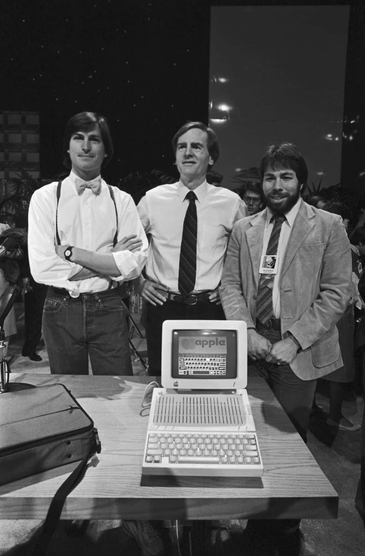  壁紙 — 三位身穿1980年代服裝的男士站在一張展示早期Apple手提電腦的桌子後面，出席科技活動。