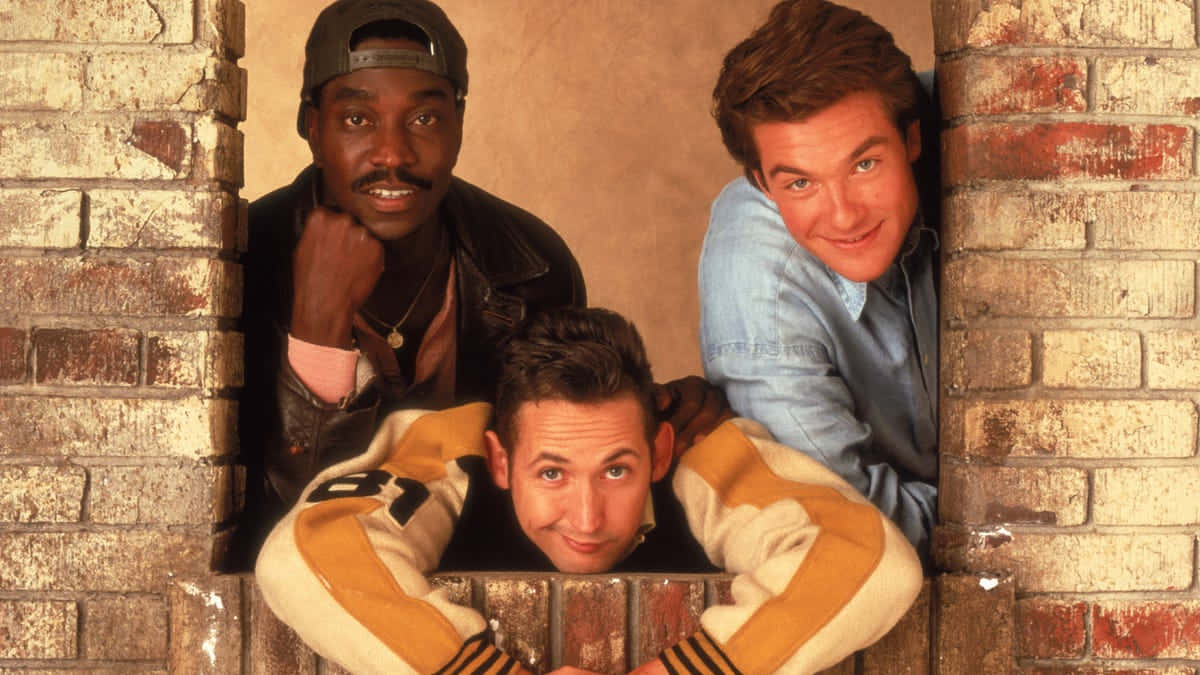  壁紙 — 三個男人透過磚牆開口窺視，表情頑皮，穿著休閒的90年代街頭服飾。