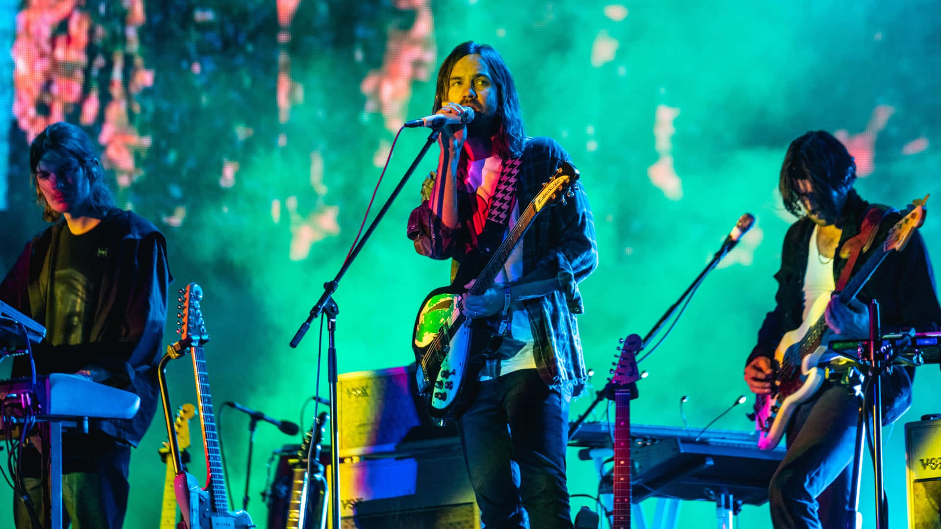  壁紙 — 三位音樂家在舞台上現場表演，沐浴在充滿活力的綠色和粉紅色舞台燈光及煙霧效果中。