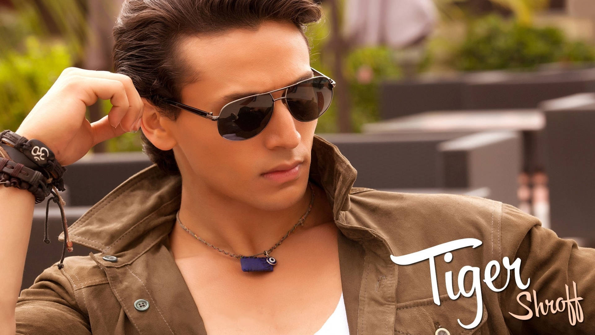 年輕的 Tiger Shroff 壁紙 — 一位戴著飛行員太陽眼鏡和棕色外套的時尚男士，在柔和的日光下自信地於戶外擺姿勢，周圍有綠植和現代家具。