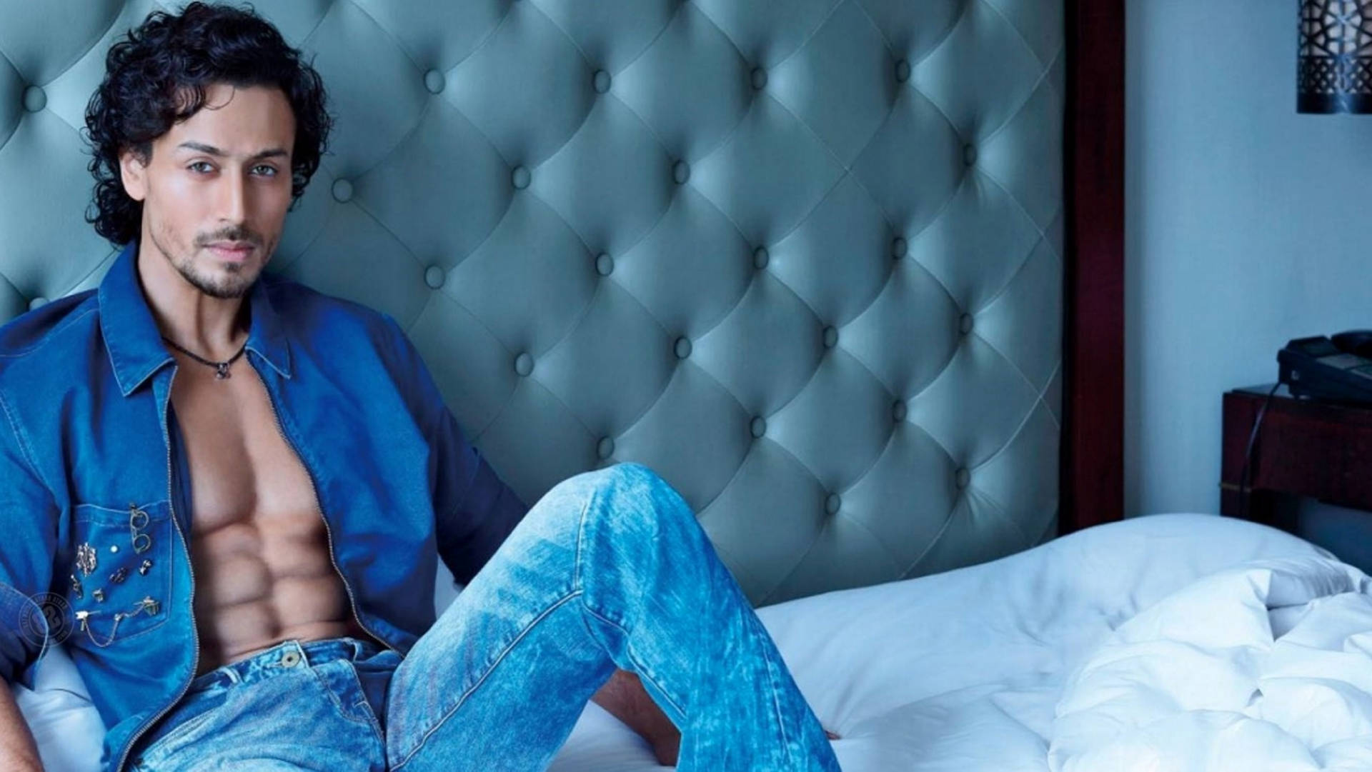 Tiger Shroff 照片拍攝 壁紙 — 一個肌肉發達的男人穿著藍色牛仔褲，在冷藍燈光下，赤裸上身躺在一張有簇絨床頭板的床上。