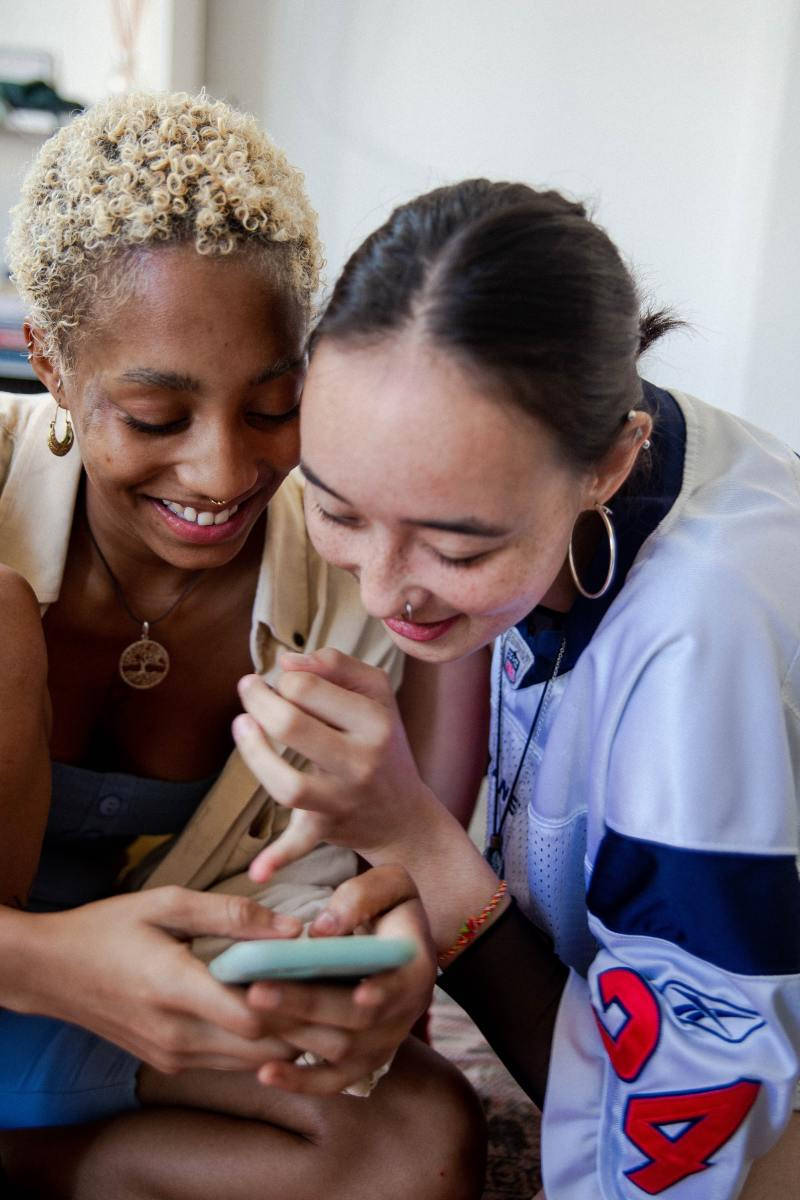 快樂朋友頭像適用於TikTok 壁紙 — 兩位微笑的女性在柔和自然光中靠近分享電話時刻。