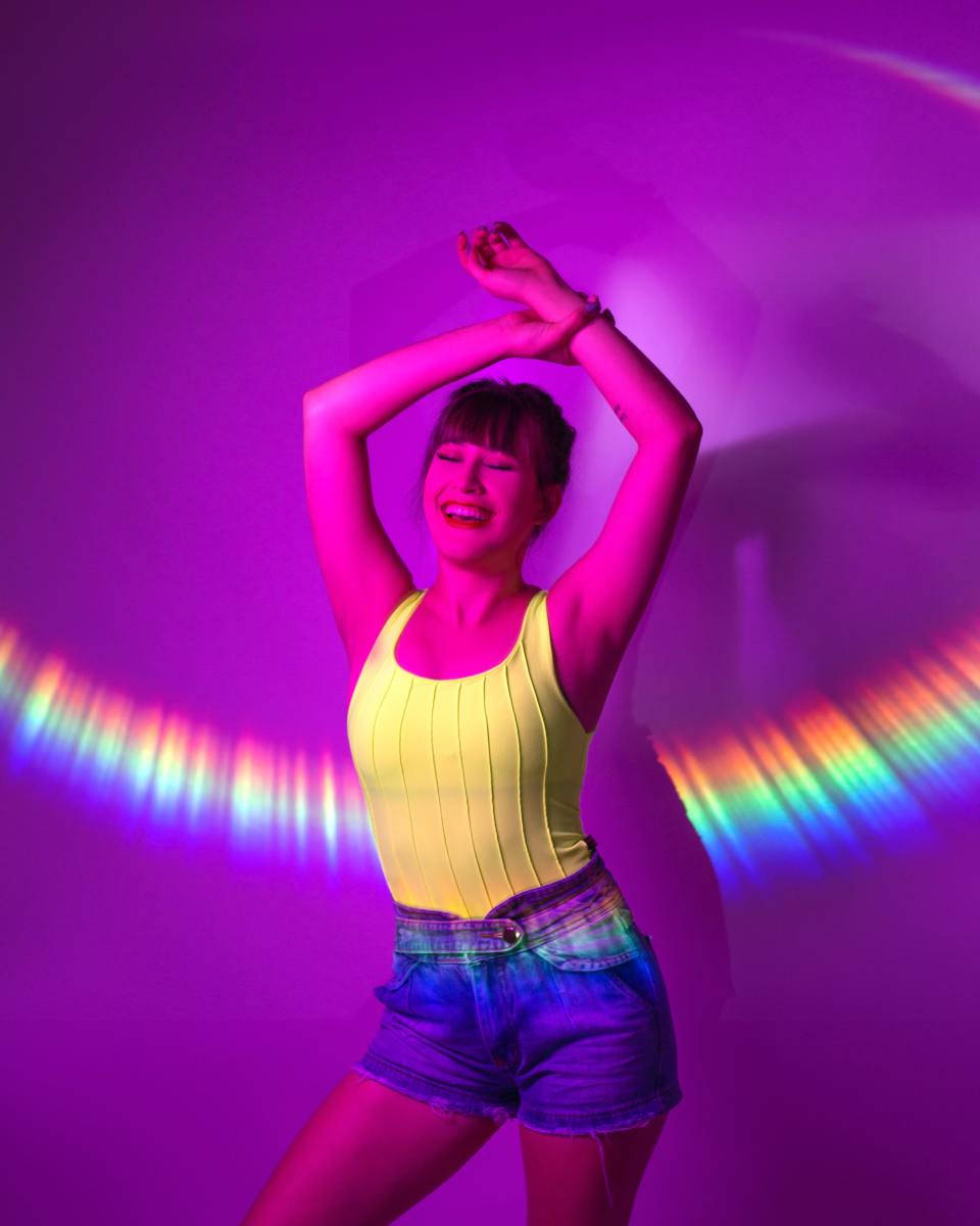 TikTok 個人頭像彩虹與快樂女孩 壁紙 — 一位身穿黃色上衣和牛仔短褲的快樂女性，在背後有彩虹弧線的鮮豔紫色燈光下跳舞。