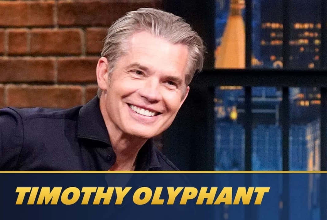 timothy olyphant 桌布 壁紙 — Timothy Olyphant 穿著深色襯衫，在磚牆和城市景觀背景下溫暖微笑，帶有金色文字疊加。