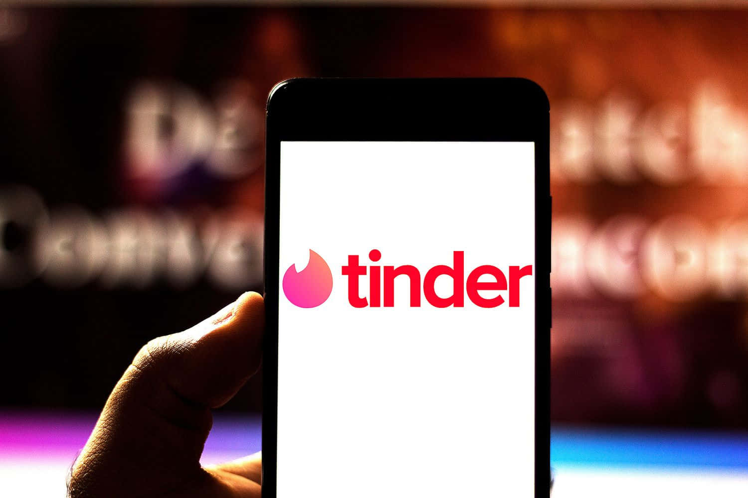 手拿著手機顯示Tinder應用程式標誌，背景模糊色彩繽紛。