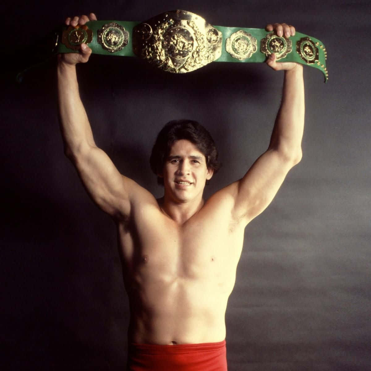 Tito Santana WWF 國際冠軍 壁紙 — 一個肌肉發達、赤裸上身的男人在黑暗的攝影棚背景下，勝利地將一條綠色和金色的冠軍腰帶高舉過頭頂。