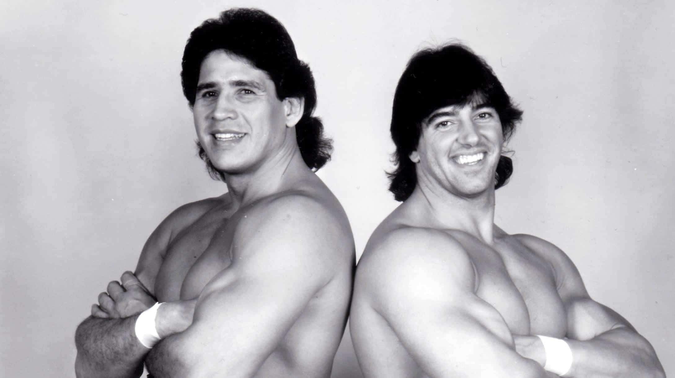 Tito Santana 與 Rick Martel 擊力組合 壁紙 — 兩位肌肉發達、頭髮長及肩膀的男士背對背站立，在黑白工作室肖像中自信地微笑。