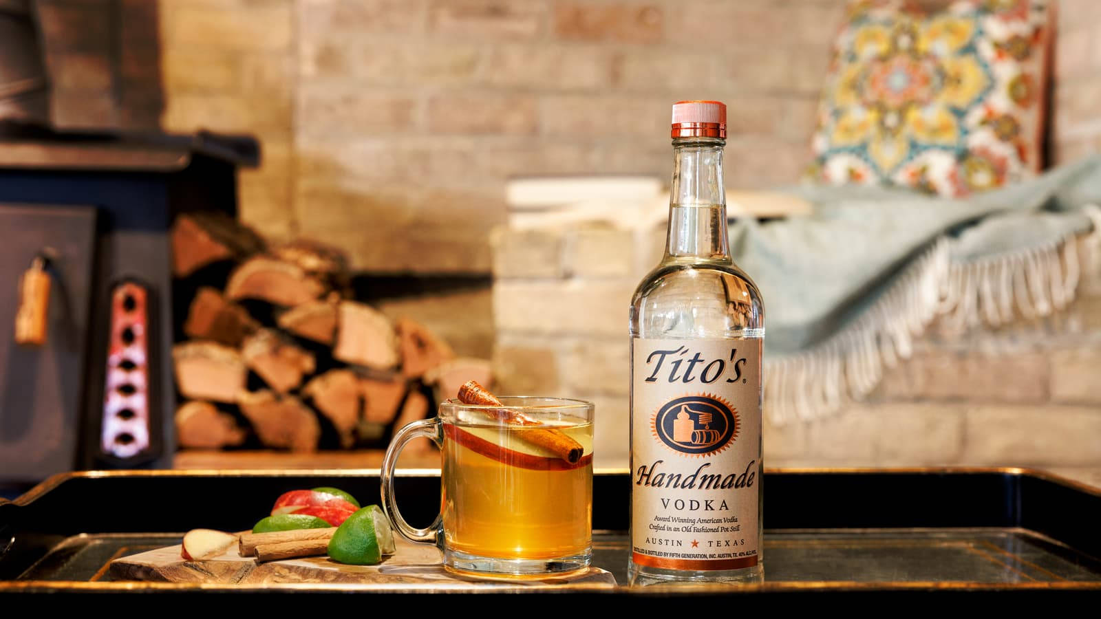 Titos伏特加配蘋果與肉桂 壁紙 — 一個舒適的冬日場景，一杯香料蘋果酒和Tito's伏特加放在壁爐旁，壁爐裡堆滿了木柴。