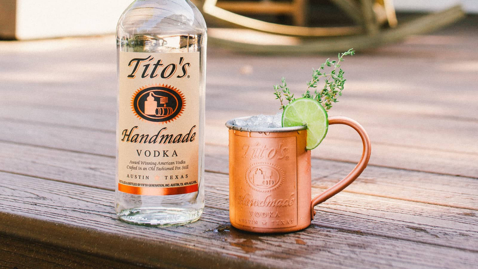 Titos Vodka 盛於銅杯中 壁紙 — 一個裝有冰塊、青檸和香草的銅杯，放在一個飽經風霜的木甲板上，旁邊是一瓶Tito's伏特加。