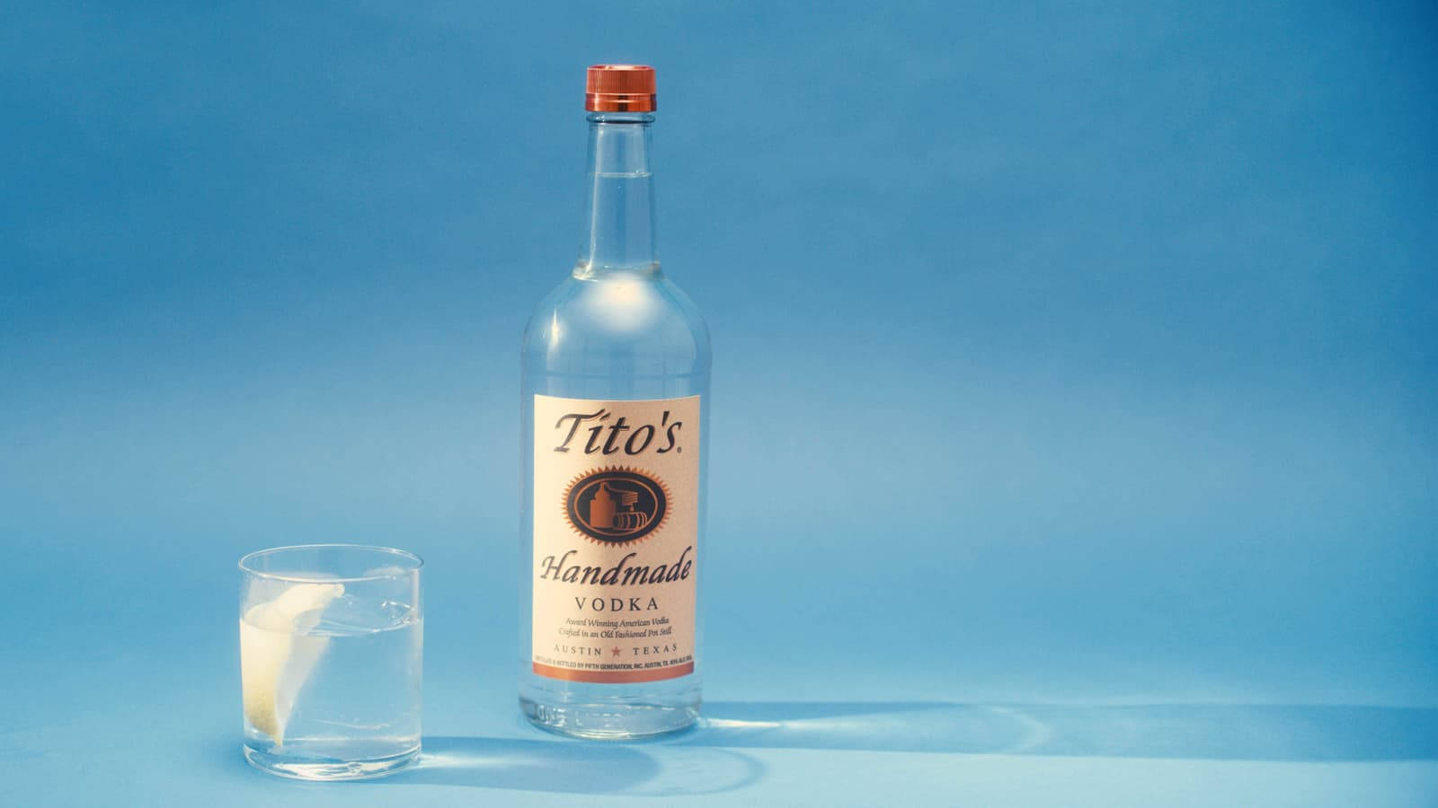 Tito's伏特加酒瓶和裝有冰塊和檸檬的玻璃杯，藍色背景