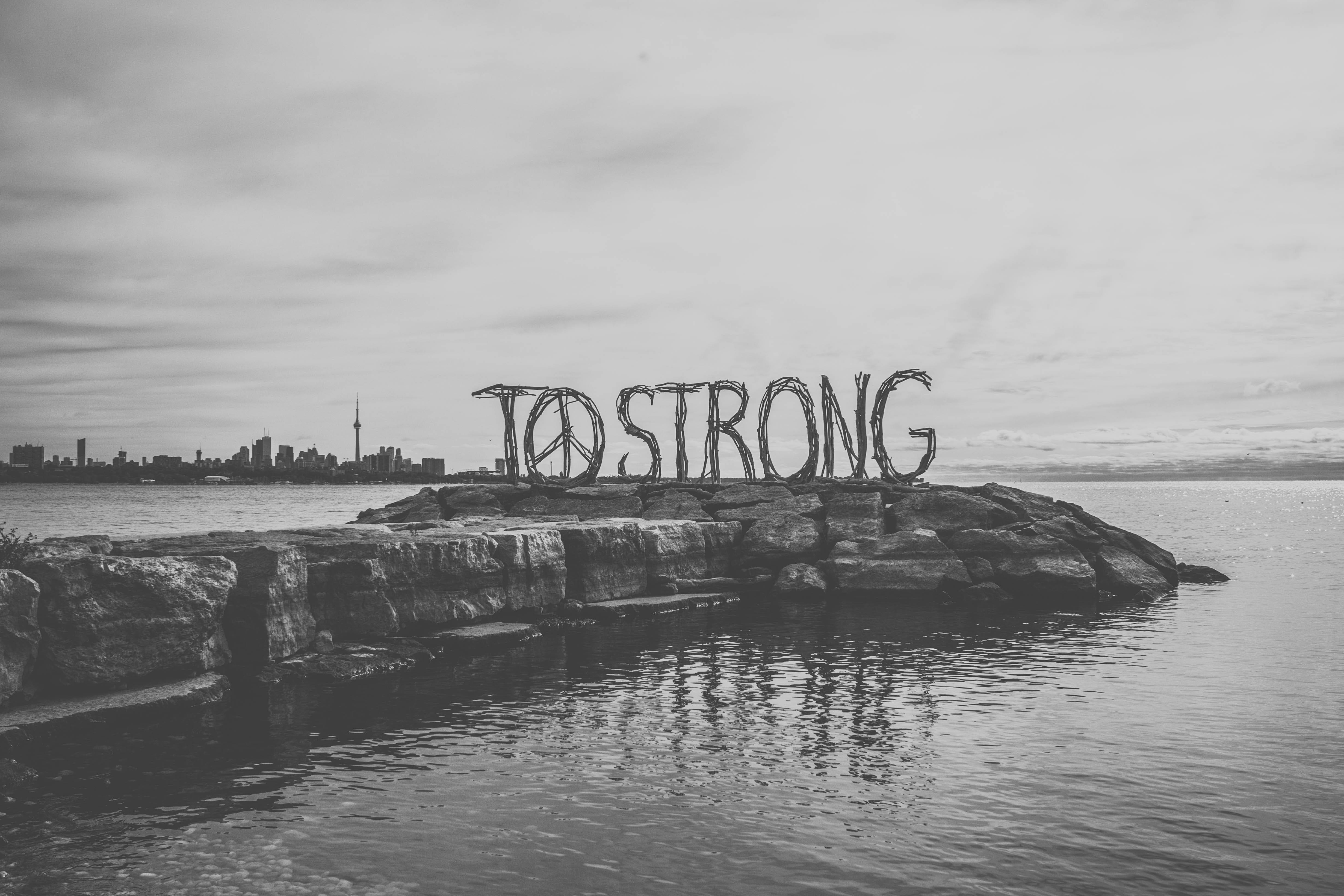 和平符號的「TO STRONG」海景 壁紙 — 岩石島上「TO STRONG」編織標誌的黑白景觀，多倫多天際線倒映在平靜的水面上。