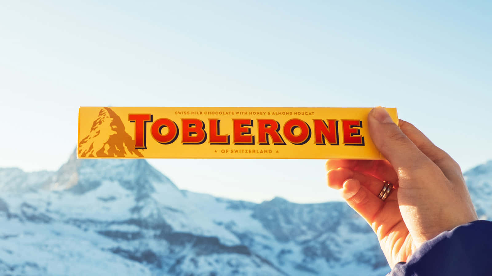 Toblerone 巧克力品牌圖標 壁紙 — 一隻手拿著 Toblerone 朱古力棒，背景是晴朗藍天下白雪覆蓋的瑞士山脈。
