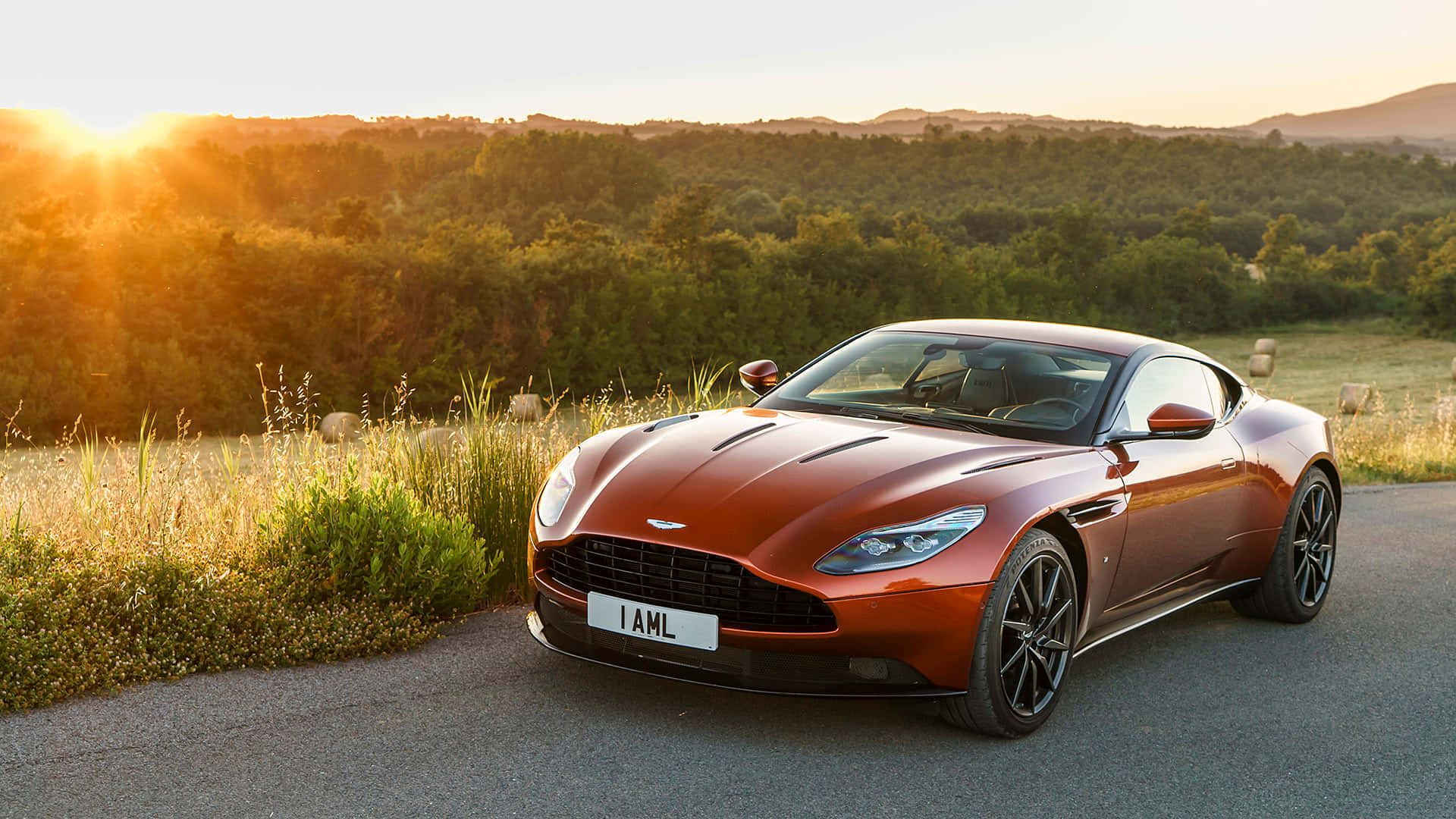 Aston Martin Db11 桌布 壁紙 — 一輛時尚的銅色Aston Martin跑車在金色夕陽下的蜿蜒道路上閃閃發光，背景是起伏的山丘和樹木。