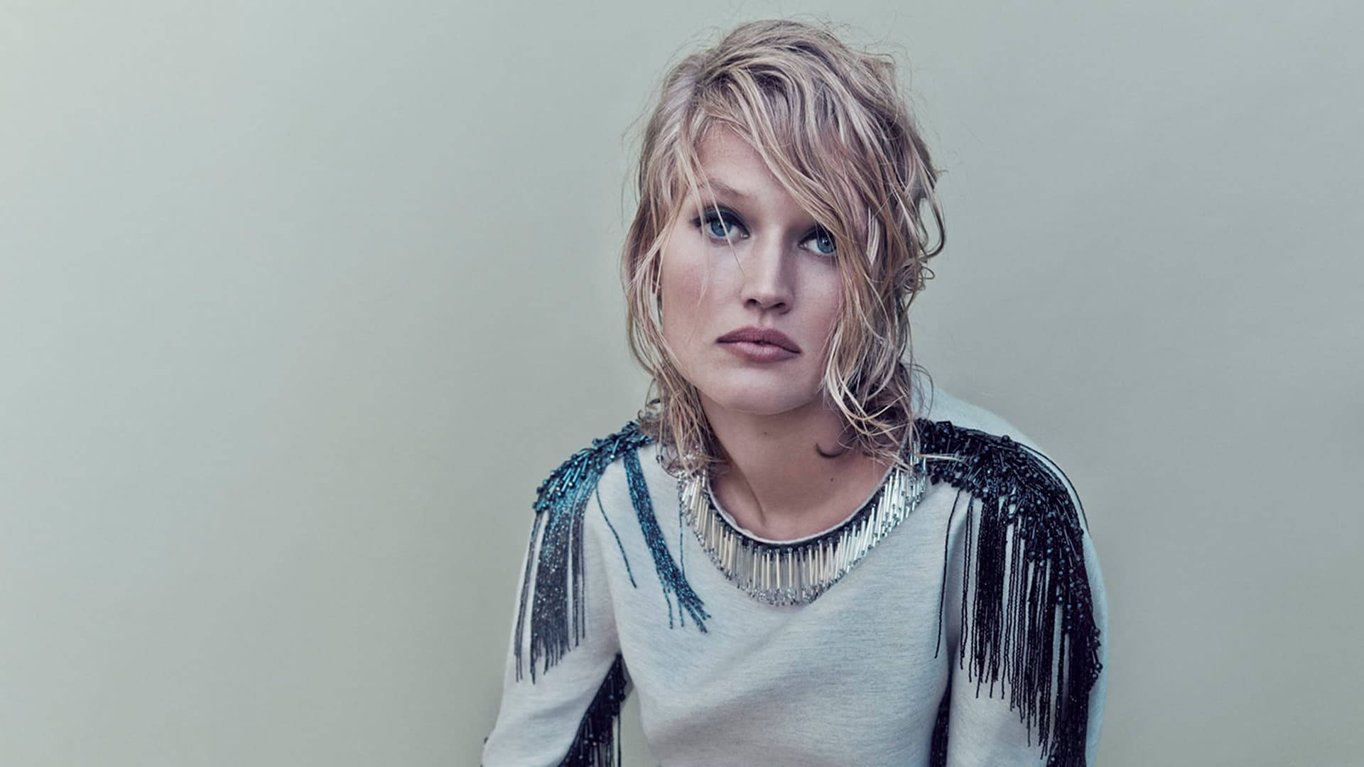 Toni Garrn 傷感表情 壁紙 — 一位頭髮蓬鬆金髮、擁有引人注目的藍眼睛的女性，穿著流蘇銀色上衣，背景是純米色牆壁。