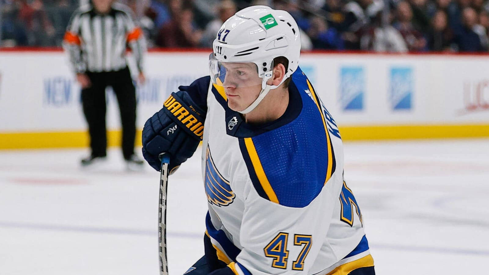 Torey Krug（2022 年 11 月） 壁紙 — 一名專注的曲棍球運動員身穿白色、藍色和金色球衣，背號47，在冰上滑行，背景有裁判。