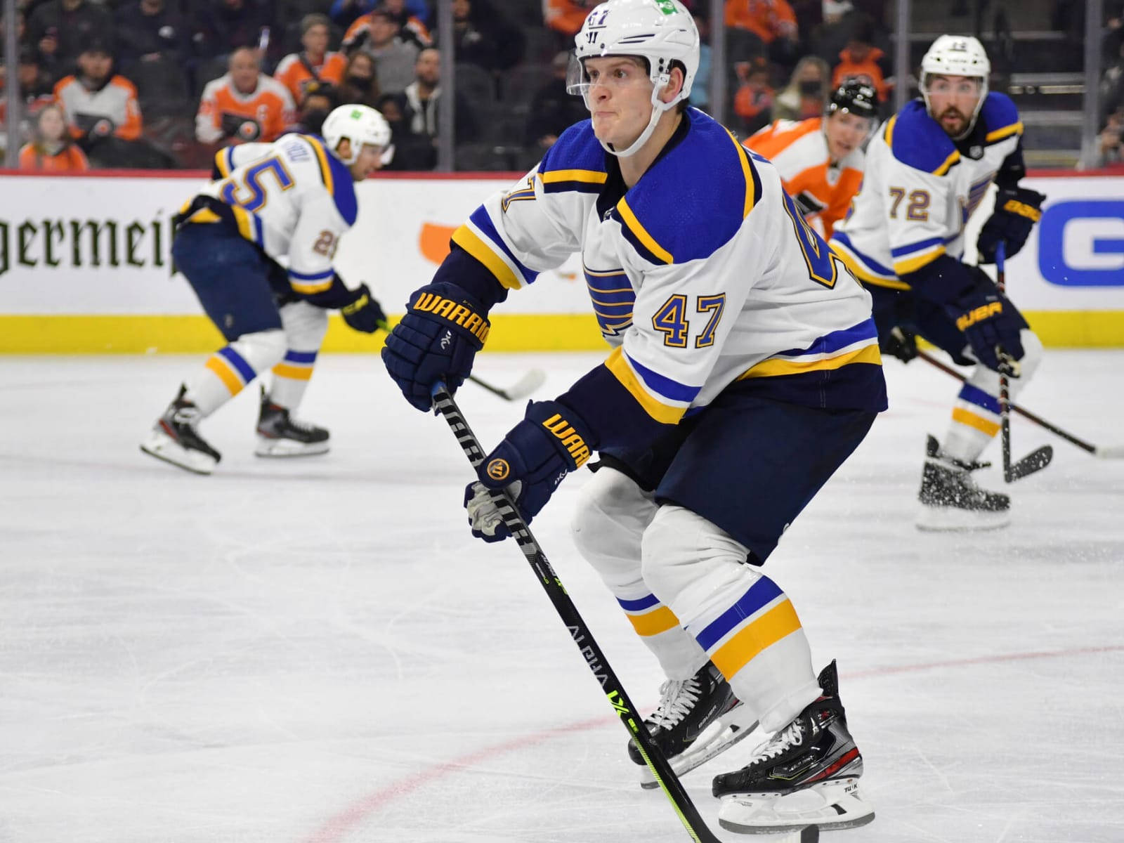 Torey Krug 與 Jordan Kyrou 及 Justin Faulk 同場 壁紙 — 一名身穿白、藍、金色球衣的冰球運動員在比賽中與隊友和對手激烈地在冰上滑行。