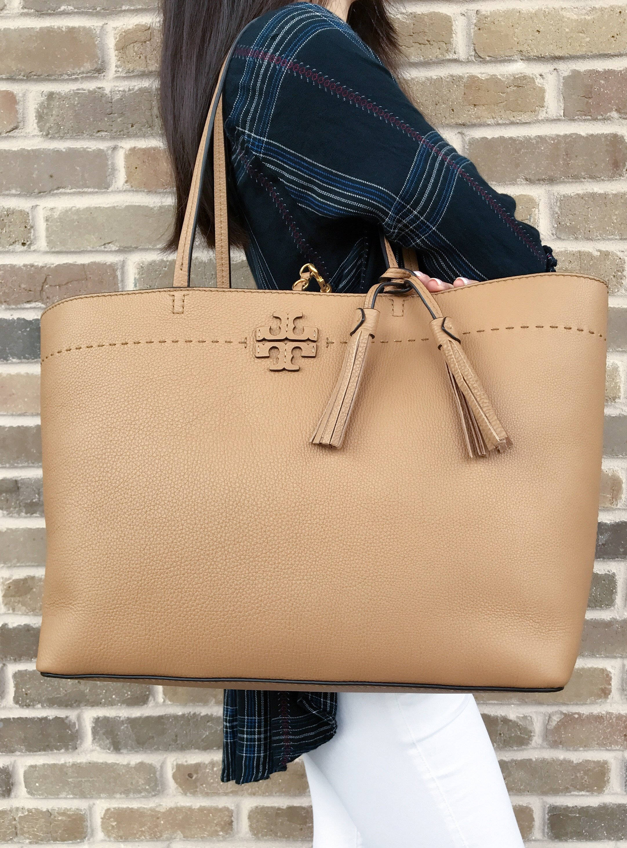 Tory Burch McGraw 手提袋 壁紙 — 一個人穿著格仔衫和白色褲子，拎著一個帶流蘇的大號棕褐色皮革托特包，背景是磚牆。